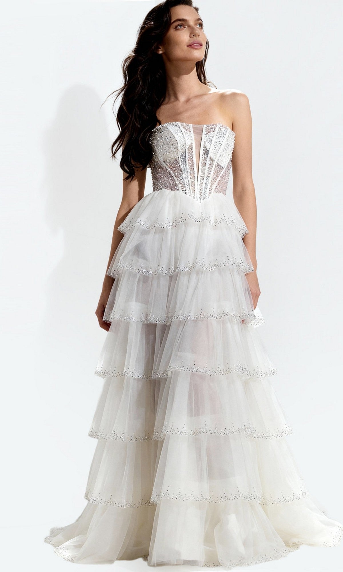 A woman stands against a plain background wearing the Jovani 47290 Long Prom Dress in off white, featuring a strapless, corset-style fitted bodice and a layered tulle, tiered skirt.